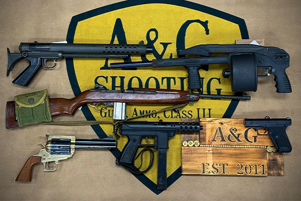 Four firearms, including rifles and handguns, displayed on a tan cloth with an A&G Shooting sign in the background.