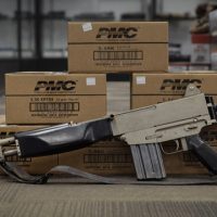 A rifle with a sling is displayed on the floor in front of stacked boxes labeled "PMC" in a store setting.