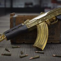 A gold-plated AK-47 rifle rests on a wooden crate, surrounded by loose rifle cartridges on the floor.