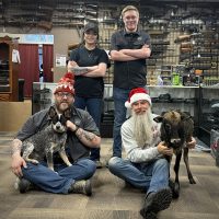 Four people pose inside a gun shop; two are seated with a dog and a goat, while two stand behind them.