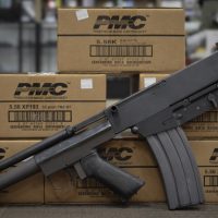 A black assault rifle is propped against stacked boxes of PMC 5.56mm centerfire rifle ammunition in a store setting.