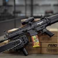 A black rifle with an attached chainsaw rests on ammunition boxes, with a box labeled "Lucky Arms" inserted in the magazine well.