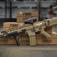 A tan FN SCAR assault rifle with scope and grip is displayed in front of PMC 5.56mm ammunition boxes on a table.
