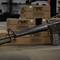 A black rifle with a bayonet attached is propped against boxes of PMC 5.56mm ammunition in a store or warehouse.