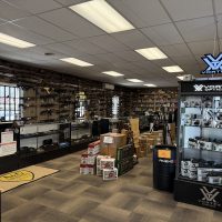 Interior of a gun store with rifles on wall racks, glass display cases, ammo boxes, and optics display.