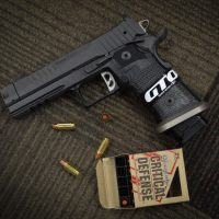 A black handgun, loose bullets, and an open box labeled “Critical Defense” are displayed on a textured carpet.