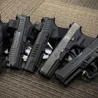 Five black semi-automatic handguns are displayed side by side on a brown patterned carpeted floor.