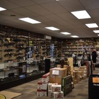 Interior of a gun shop with rifles on wall racks, glass display cases, and ammo boxes stacked on the floor.