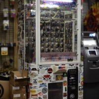 A claw machine covered in various stickers stands indoors near an ATM and some shelves with boxes and merchandise.